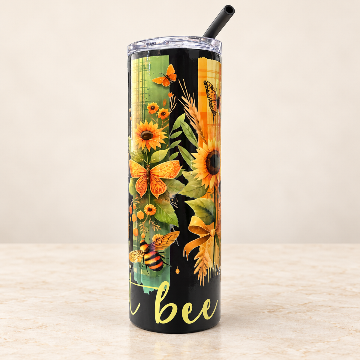 A tall insulated tumbler with a clear lid and black straw, featuring a decorative sunflower, butterfly, bee, and wheat design on a black background with yellow script text near the bottom, set on a light stone surface against a beige wall.
