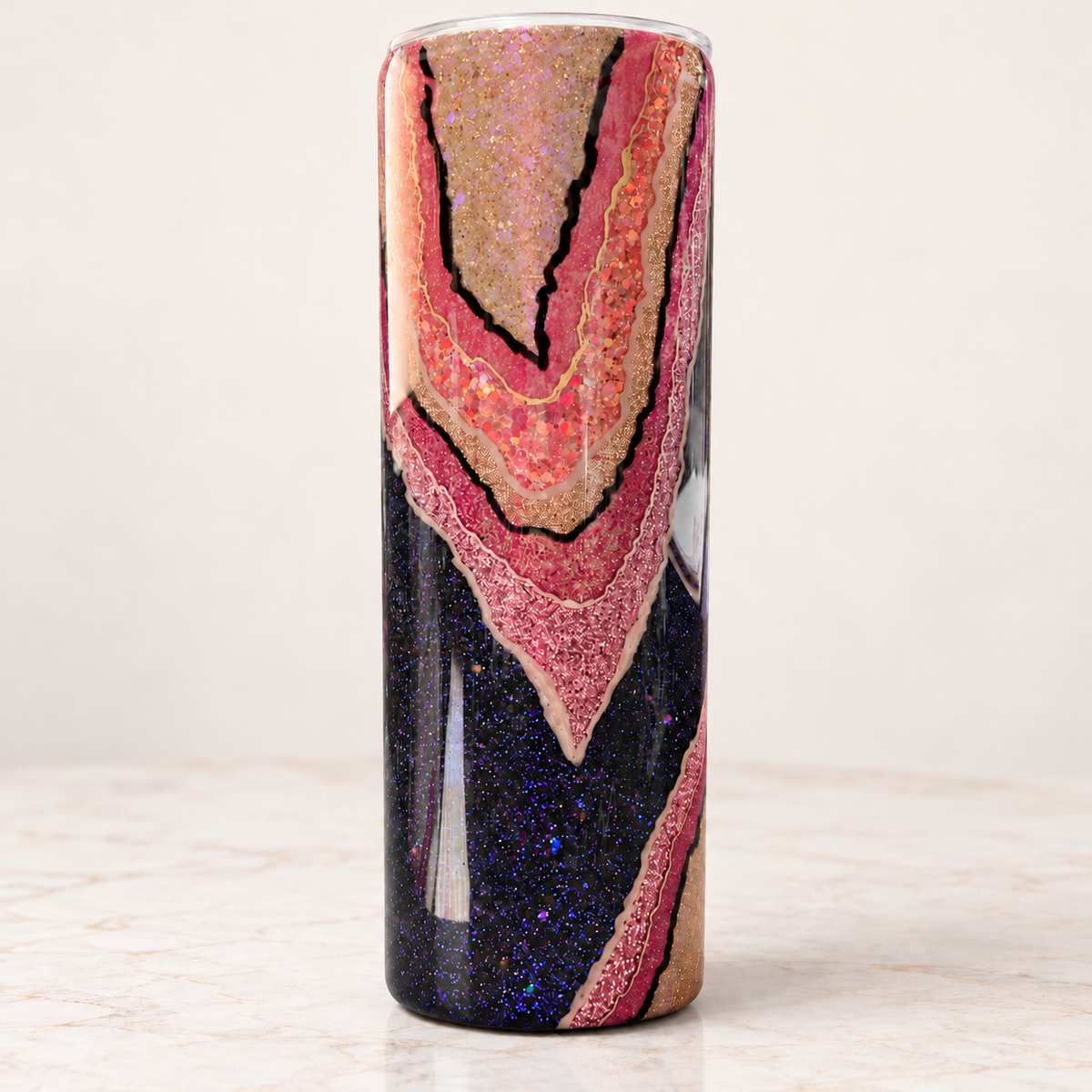 A tall cylindrical tumbler with a glittery geode pattern featuring pink, gold, and dark blue sections, shown on a light marble surface against a neutral background.