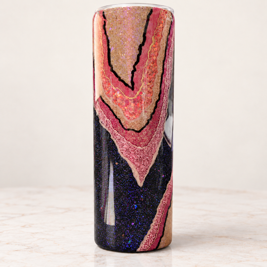 A tall cylindrical tumbler with a glittery geode pattern featuring pink, gold, and dark blue sections, shown on a light marble surface against a neutral background.