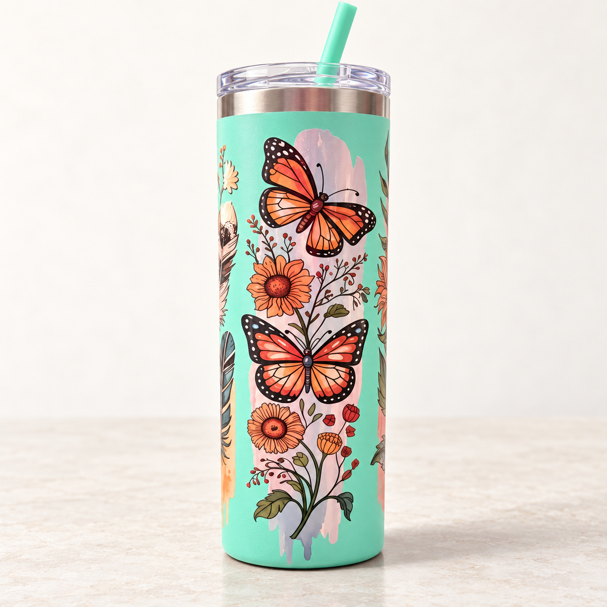 A tall insulated tumbler with a clear lid and mint green straw, decorated with orange monarch butterflies, flowers, and leaves on a mint green background, set on a light surface against a white wall.