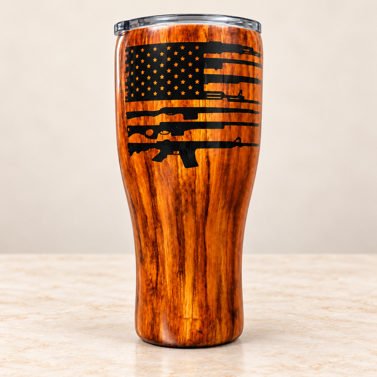 A tall insulated tumbler with a wood-grain finish and a clear lid, featuring a black American flag design with rifle silhouettes on the front, set on a light stone surface against a beige background.