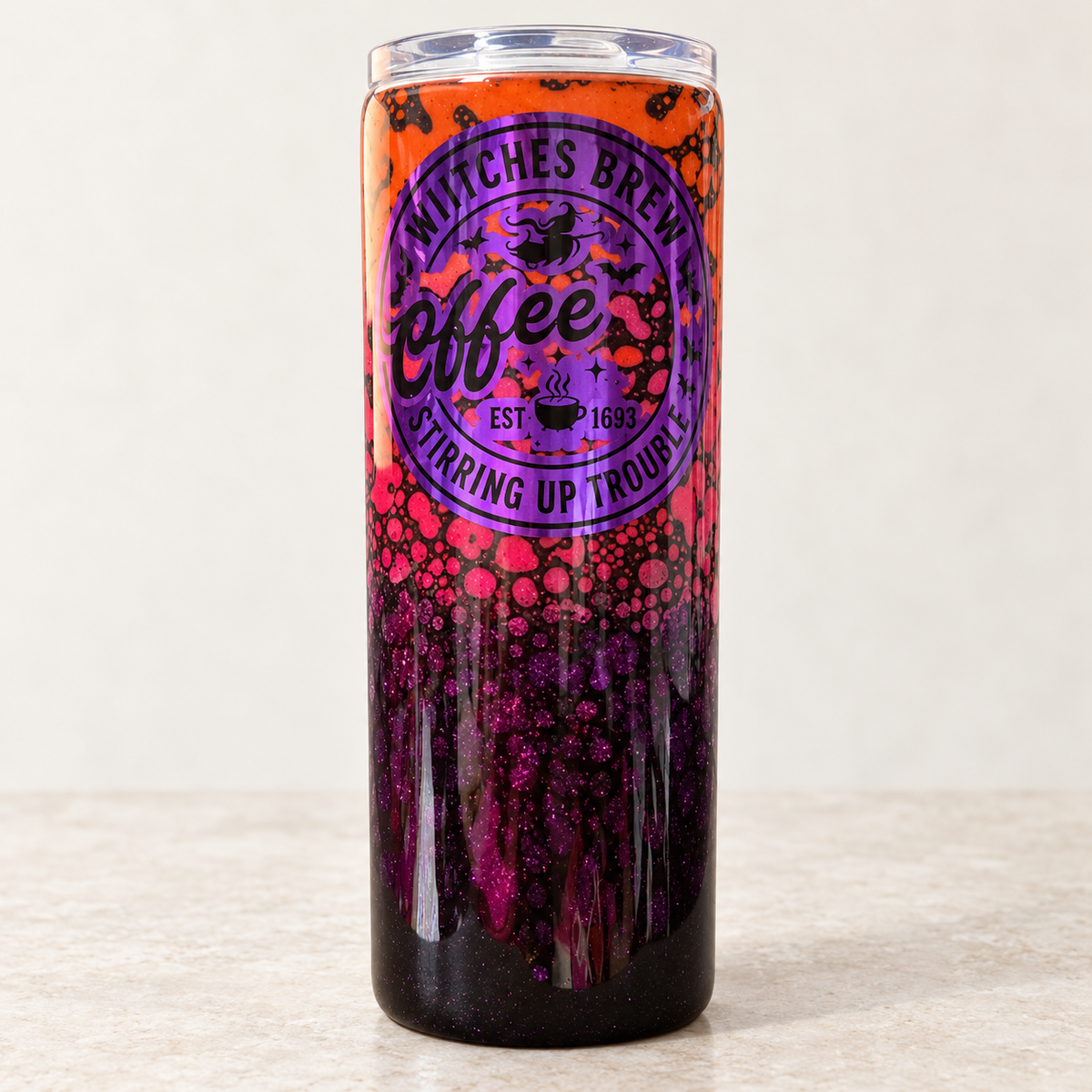 A tall insulated tumbler with a clear lid, featuring a Halloween-themed design with orange at the top, a purple circular logo reading 'Witches Brew Coffee', and dark glittery purple splatter patterns near the bottom, set against a neutral beige background.