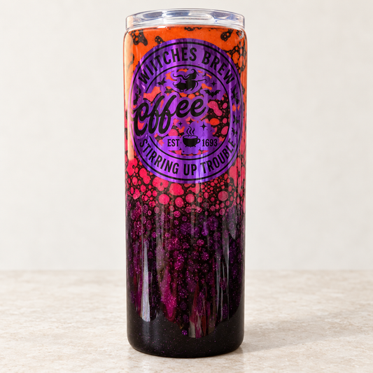 A tall insulated tumbler with a clear lid, featuring a Halloween-themed design with orange at the top, a purple circular logo reading 'Witches Brew Coffee', and dark glittery purple splatter patterns near the bottom, set against a neutral beige background.
