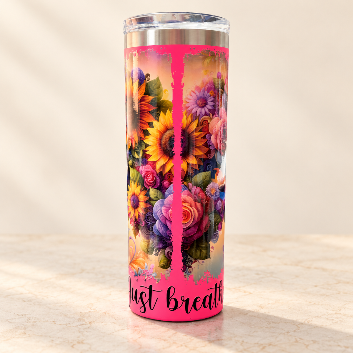 A tall insulated tumbler with a clear lid, featuring a bright pink base and a colorful floral print with sunflowers, roses, and purple blossoms. The bottom of the tumbler has the phrase 'Just breathe' in black script.