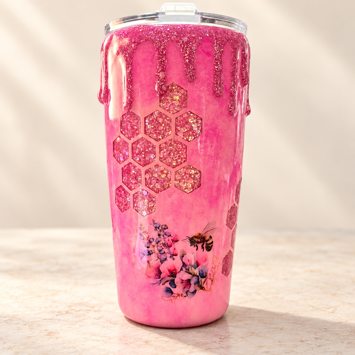 A pink glitter tumbler with a clear lid, decorated with a honeycomb pattern, dripping pink glitter, a bee, and floral artwork, set on a light stone surface.