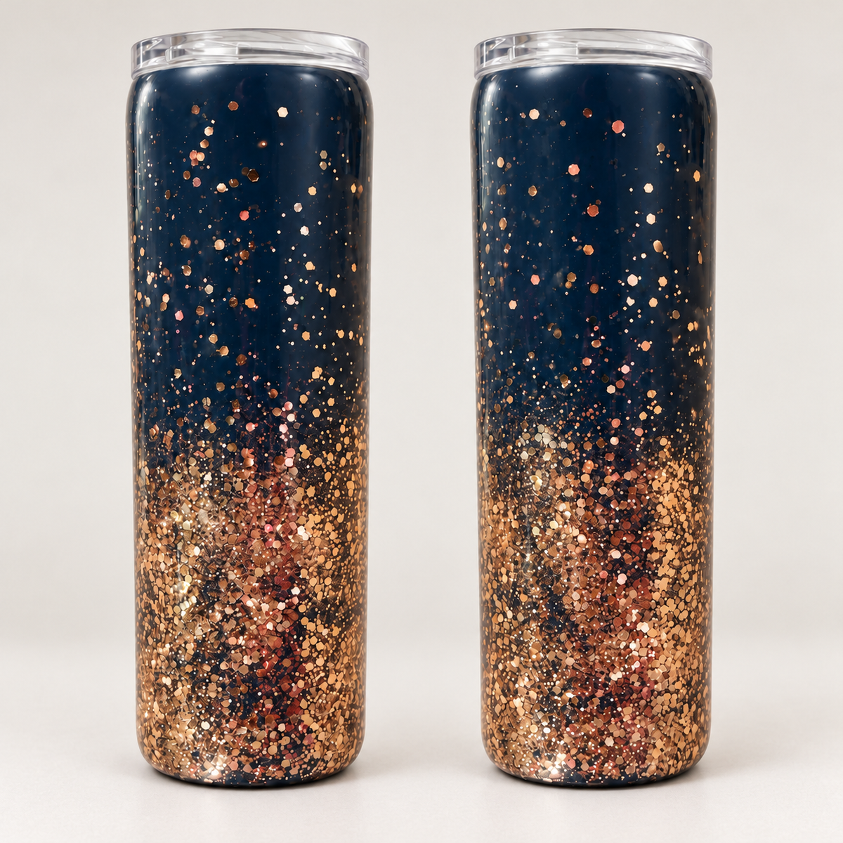 Two tall skinny tumblers with clear lids, decorated with a navy blue top half and dense rose gold glitter bottom half, shown side-by-side on a light gray background.