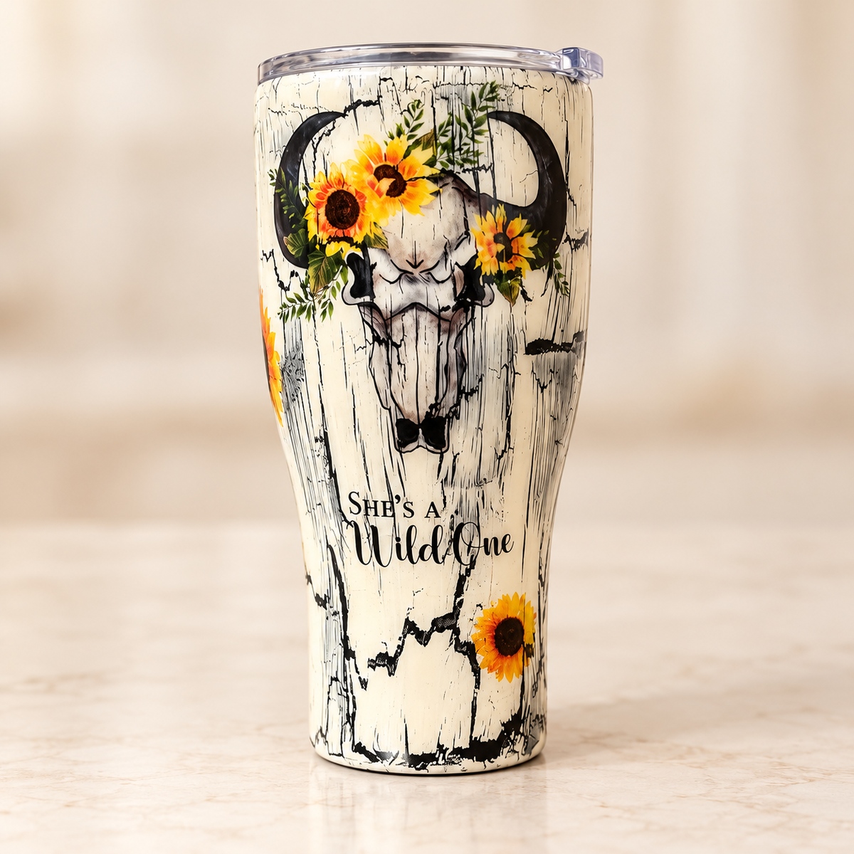A cream-colored insulated tumbler with a rustic wood-grain crackle pattern, featuring a central bull skull design decorated with yellow sunflowers and green leaves. The text 'She's a Wild One' is printed near the bottom.