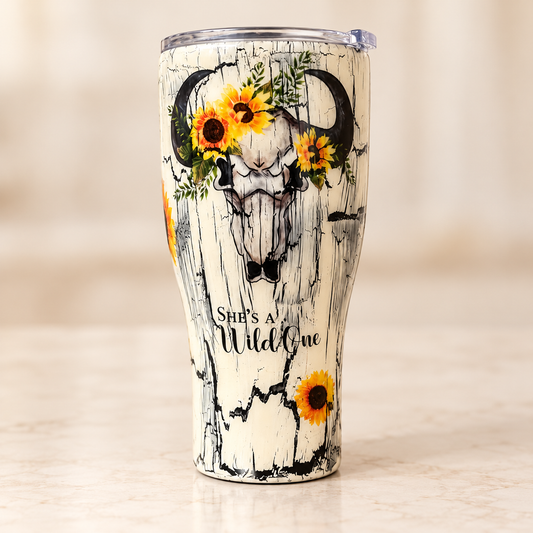A cream-colored insulated tumbler with a rustic wood-grain crackle pattern, featuring a central bull skull design decorated with yellow sunflowers and green leaves. The text 'She's a Wild One' is printed near the bottom.