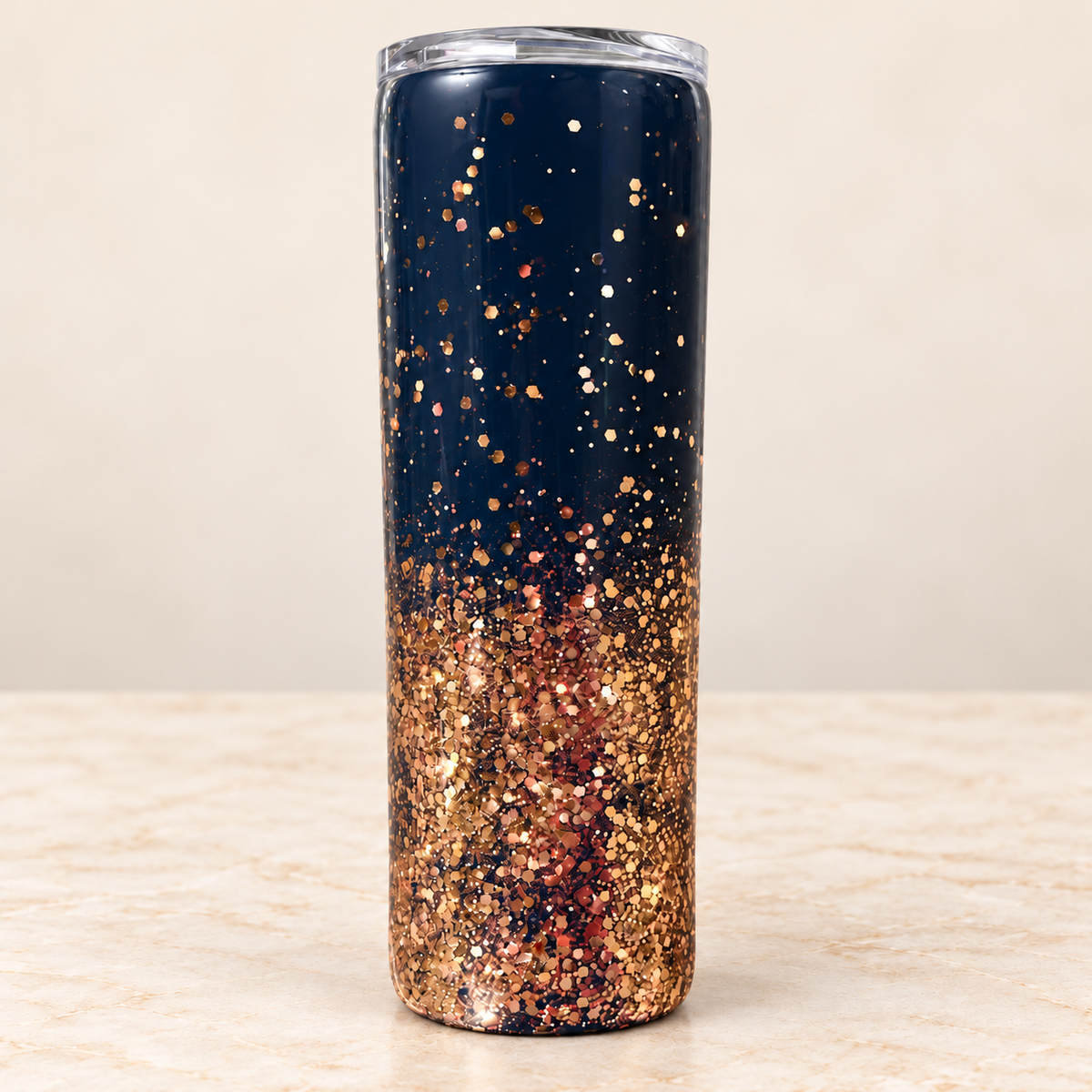 A tall, skinny insulated tumbler with a clear lid, featuring a navy blue top half and a rose gold glitter bottom half, set against a neutral beige background.