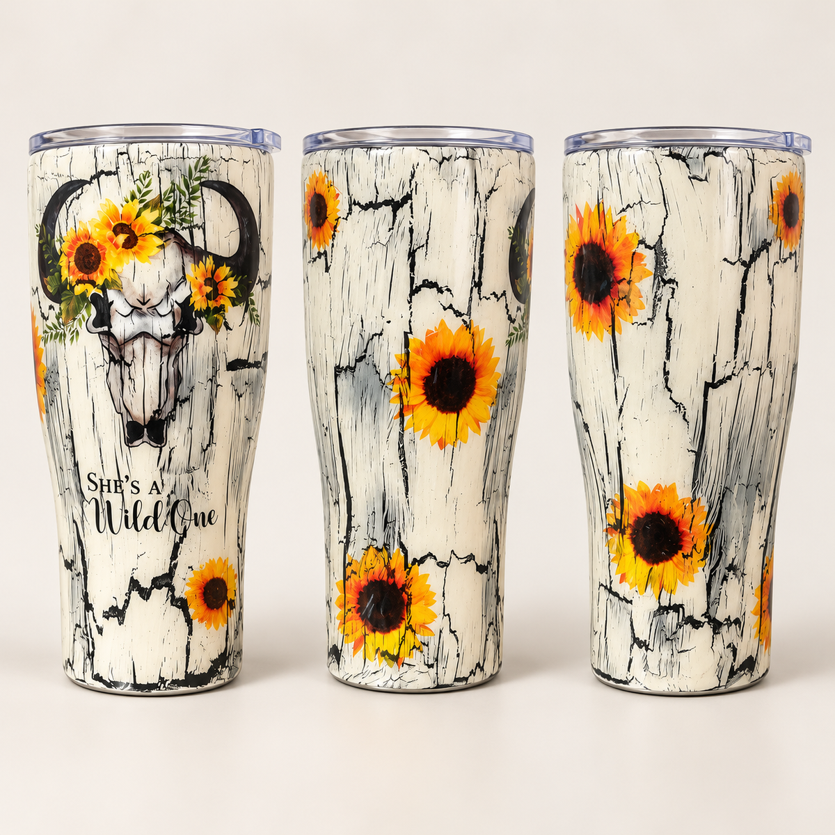 Three views of a insulated tumbler with a cracked white wood-style print, yellow sunflowers, a bull skull graphic, and the phrase 'She's a Wild One' on a light neutral background.