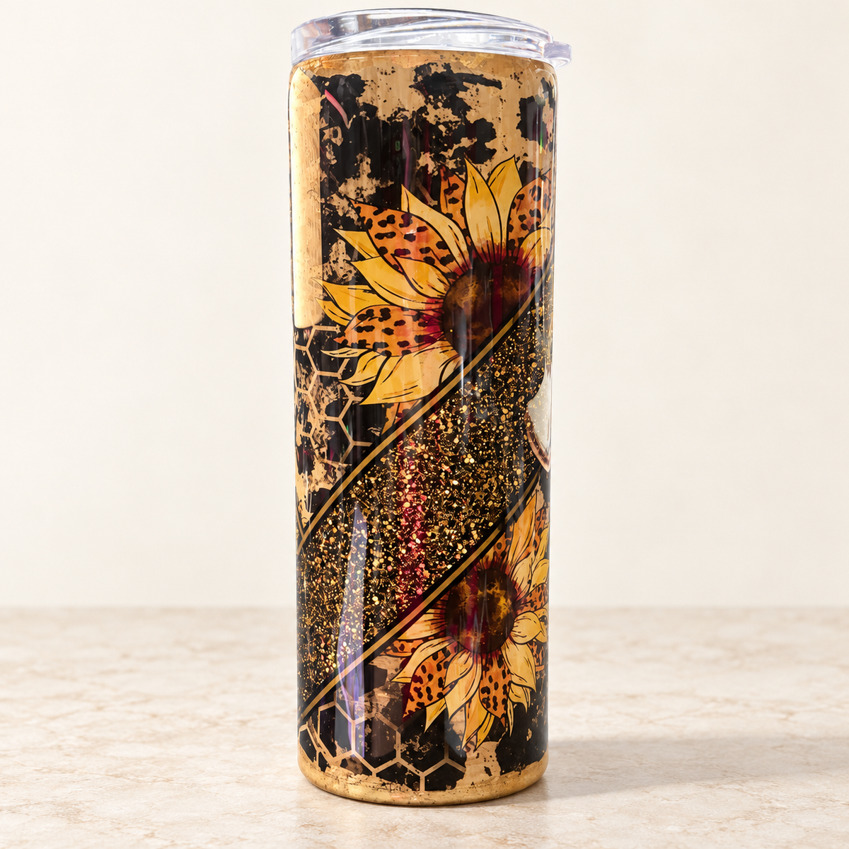 A tall insulated tumbler with a clear lid, featuring a printed design of two yellow sunflowers, gold glitter, and a black honeycomb pattern on a light beige background.