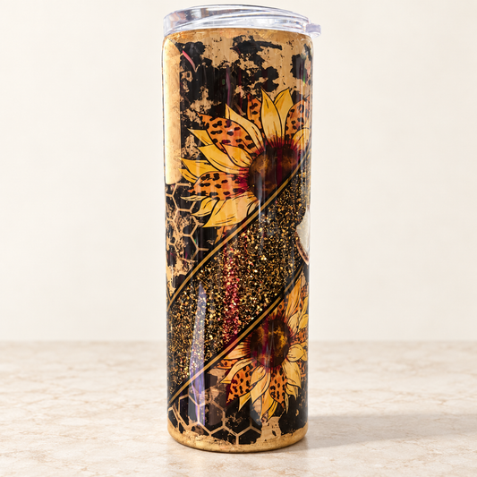 A tall insulated tumbler with a clear lid, featuring a printed design of two yellow sunflowers, gold glitter, and a black honeycomb pattern on a light beige background.
