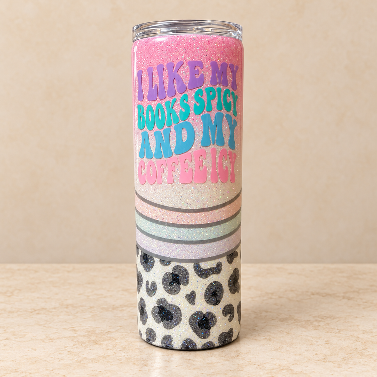 A tall insulated tumbler with a clear lid, featuring a glittery pink and white body with the text 'I LIKE MY BOOKS SPICY AND MY COFFEE ICY' in colorful lettering, plus a black-and-cream leopard print section near the bottom, set against a beige background.