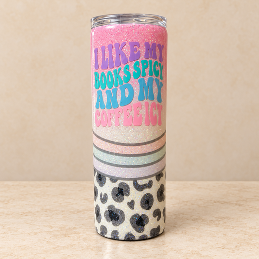 A tall insulated tumbler with a clear lid, featuring a glittery pink and white body with the text 'I LIKE MY BOOKS SPICY AND MY COFFEE ICY' in colorful lettering, plus a black-and-cream leopard print section near the bottom, set against a beige background.